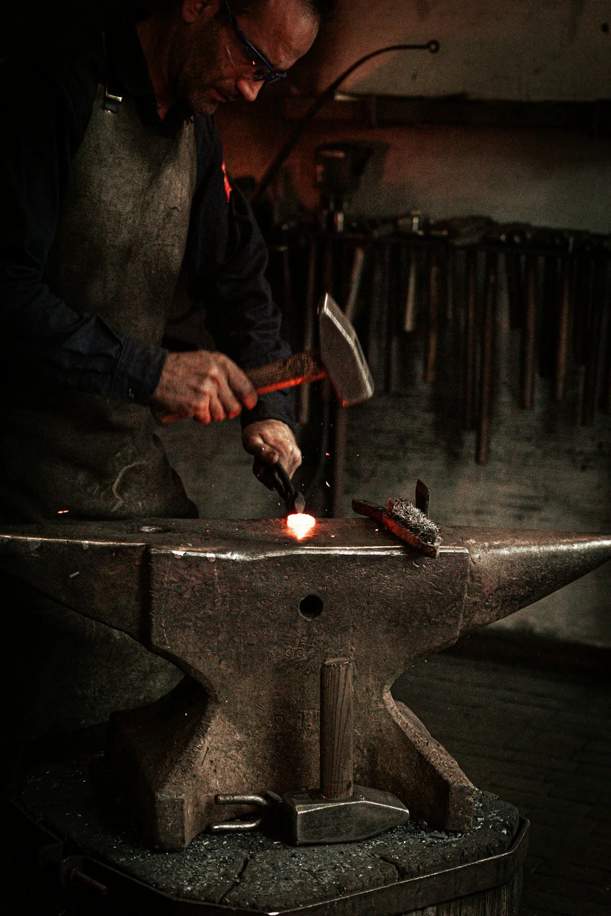 Nahaufnahme der Hände eines Meisterhandwerkers bei der Metallbearbeitung neben einem historischen Schwarzweißfoto einer traditionellen Schmiede